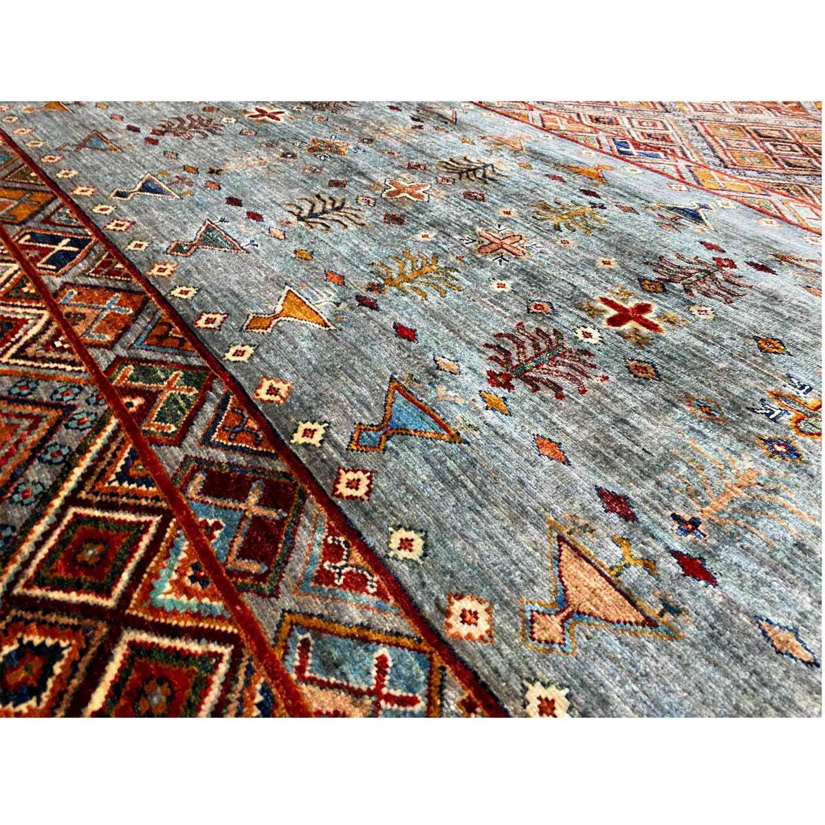Nahaufnahme: graublauer Wollteppich mit bunten Kreuz- und Ornamenten, rechteckiger Bordüre